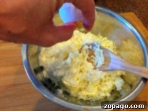 A hand cracks an egg into a metal bowl containing a mixture of shredded cheese and cream, with a wooden spoon in the bowl, preparing the filling for an easy crockpot lasagna recipe.