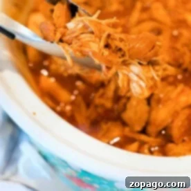 Close-up of pulled chicken being held by tongs above a crockpot. This BBQ chicken recipe showcases tender, shredded meat in a savory sauce, with the slow cooker lined with a floral pattern on the outside.