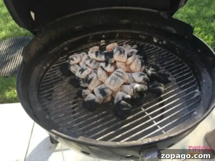 Charcoal briquettes heating up for beer can chicken on a charcoal grill, ensuring proper indirect heat for juicy results. #charcoalgrill #beercanchicken A round charcoal grill with its lid open, showing a pile of hot, partially burned charcoal briquettes in the center, ready to be spread for indirect heat cooking.