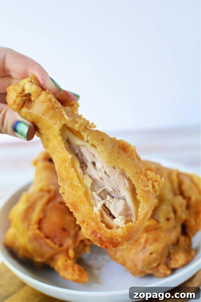 Crispy Skillet Chicken 2 A hand holds a crispy pan-fried chicken drumstick with a bite taken out, exposing the juicy inside; plate in background.