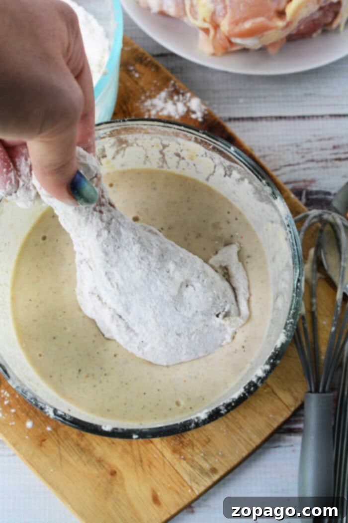Crispy Skillet Chicken 3 A hand dips a floured chicken drumstick into a bowl of batter on a wooden cutting board, preparing it for delicious pan fried chicken.