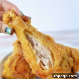 A hand holds a crispy pan fried chicken drumstick with a bite taken out, exposing the juicy inside; plate in background.