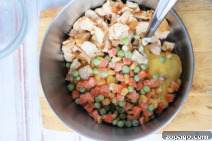 A metal bowl containing a vibrant mix of chopped rotisserie chicken, colorful frozen mixed vegetables, creamy condensed soup, and a measuring cup of Bisquick, set on a rustic wooden cutting board for easy preparation of Chicken and Dumplings.