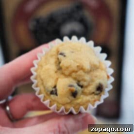 A hand holding a Kodiak banana chocolate chip muffin in a white paper liner, with more banana muffins and a box in the background.