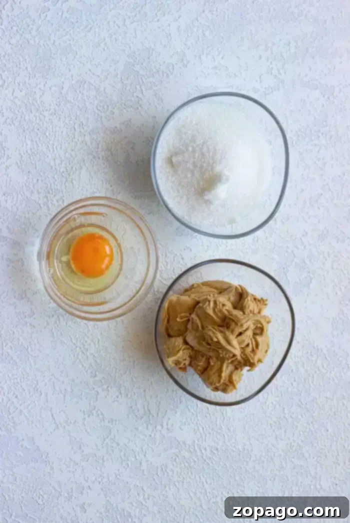 Golden Peanut Butter Thumbprints 3 A top-down view of three bowls: raw egg, granulated sugar, and creamy peanut butter – essential for 3-ingredient cookies.