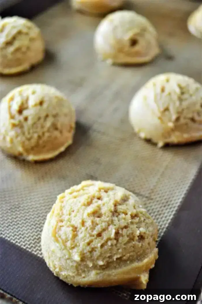 Classic Peanut Butter Thumbprint Cookies 6 Scoops of unbaked Peanut Butter and Grape Jelly cookie dough are placed on a baking sheet covered with a silicone mat. The dough appears slightly textured and is evenly spaced apart, with some of the scoops in the foreground and others in the background.