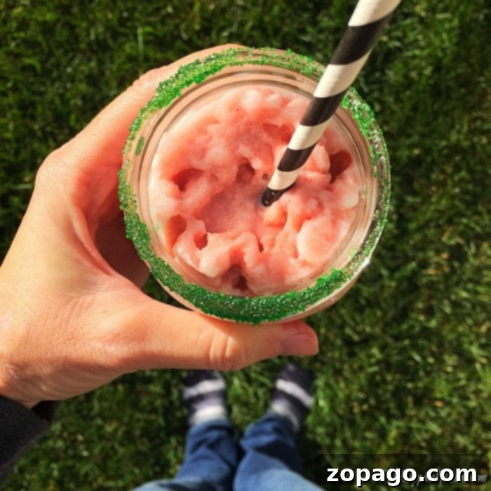 How to make a peppermint frappuccino at home with just 3 ingredients! Easy recipe to make a copycat Starbucks frappuccino at home. A person enjoys a glass of vibrant pink peppermint frappuccino with a black and white striped straw, its rim beautifully dusted in green sugar. Standing casually on the grass in socks and blue jeans, the image evokes a delightful moment, inspiring anyone to discover how to make a peppermint frappuccino for an equally delightful experience.