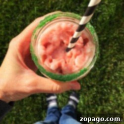 A person enjoys a glass of pink slushy with a black and white striped straw, its rim dusted in green sugar. As they stand on the grass in socks and blue jeans, you might wonder how to make a peppermint frappuccino for an equally delightful treat.