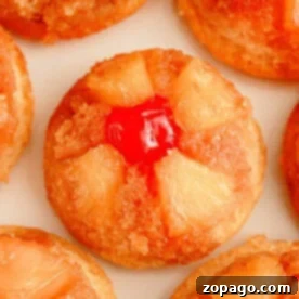 Close-up of individual pineapple upside-down cupcakes. Each cupcake showcases pineapple chunks arranged in a circular pattern, crowned with a bright red cherry at the center. The cupcakes boast a golden-brown caramelized surface.