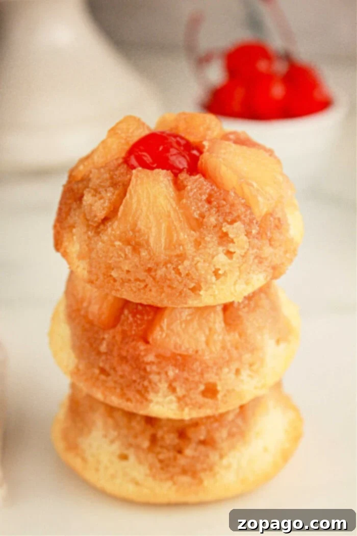 Easy Cake Mix Pineapple Upside Down Cupcakes 8 Three mini pineapple upside-down cupcakes are stacked on top of each other. Each cupcake features caramelized pineapple slices and a maraschino cherry on top. A small bowl of cherries is blurred in the background.