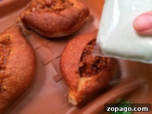 Close-up of a hand pouring white sauce from a bag onto three baked sloppy joe bread bowls filled with cooked minced meat on a brown tray.