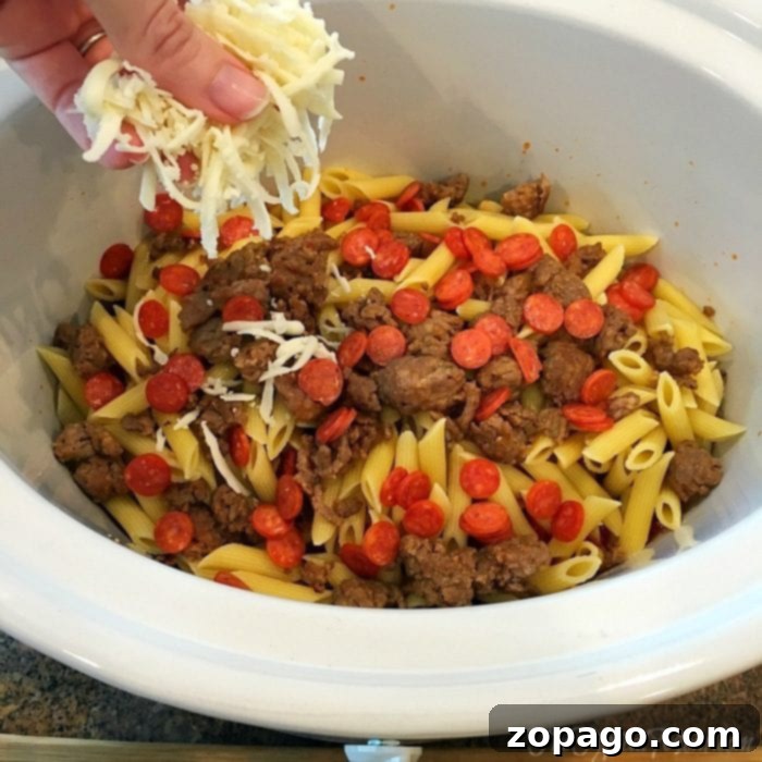 This slow cooker pizza casserole recipe is amazing! So easy to make in your crockpot and great for dinner or a party with kids. #slowcooker #pizza #crockpot #casserole #crockpotrecipes #thetypicalmom A hand sprinkles cheese over a slow cooker pizza casserole with penne, sausage, and pepperoni.