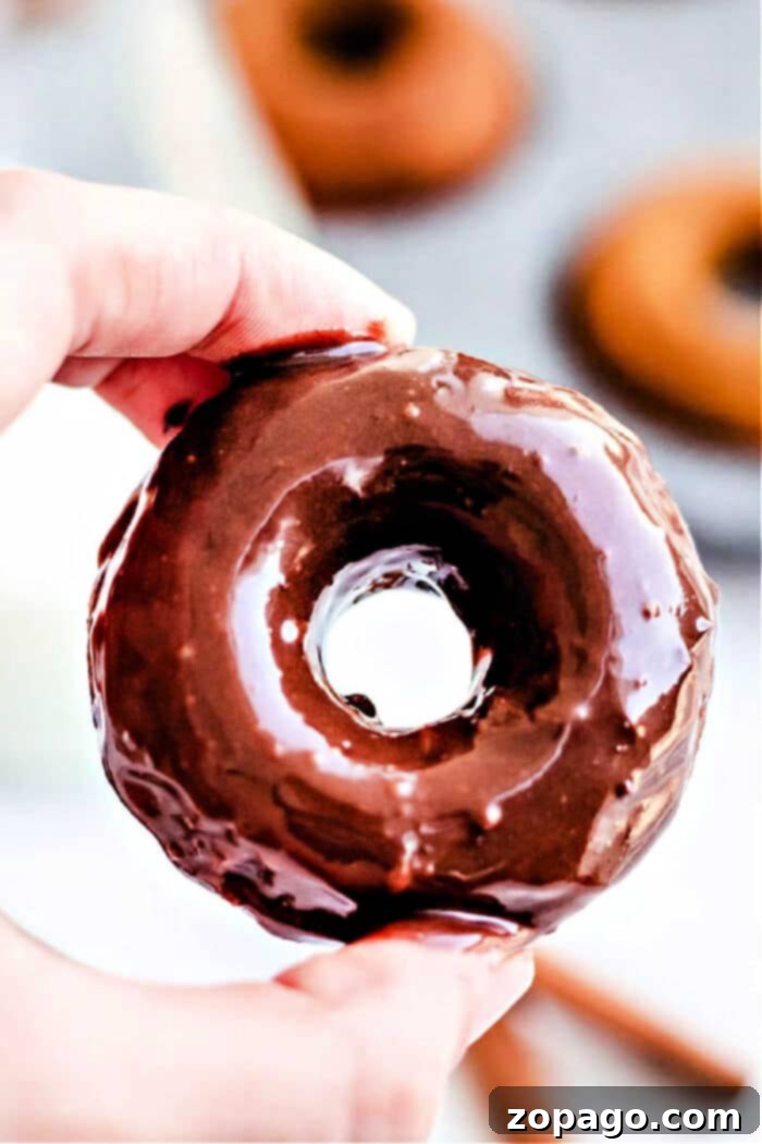 Pumpkin Spice Baked Donuts 2 Close-up of a hand holding a glazed donut with blurred baked pumpkin donuts in the background, showcasing their delicious appeal.