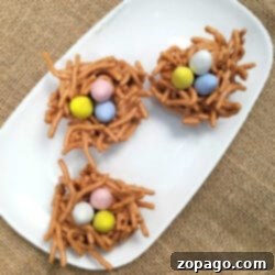 Three butterscotch haystacks shaped into cookie nests, made of pretzel sticks and filled with colorful candy eggs, sit on a white plate.