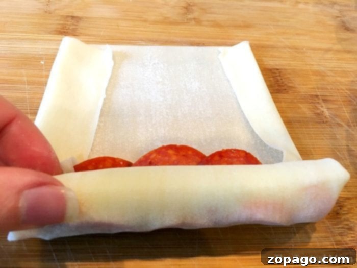 Crispy Pizza Bites 5 A hand demonstrating how to roll an egg roll wrapper with pepperoni and cheese on a wooden surface.