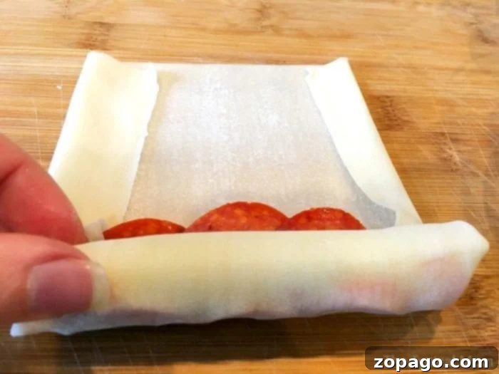 Crispy Pizza Bites 5 A close-up of hands expertly rolling an egg roll wrapper filled with pepperoni and cheese on a rustic wooden cutting board, showcasing the meticulous preparation of delicious pizza egg rolls.
