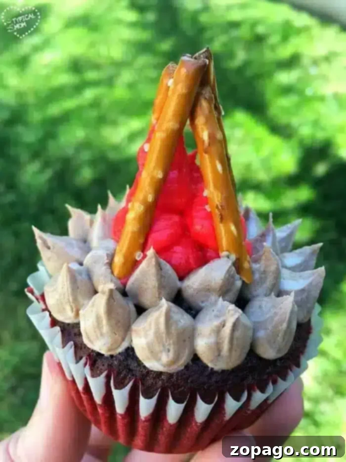 Happy Camper Cupcakes 6 Hand holding a perfectly crafted camping cupcake with vibrant red flames and sturdy pretzel logs, set against a blurred grassy background, evoking an outdoor camping scene.
