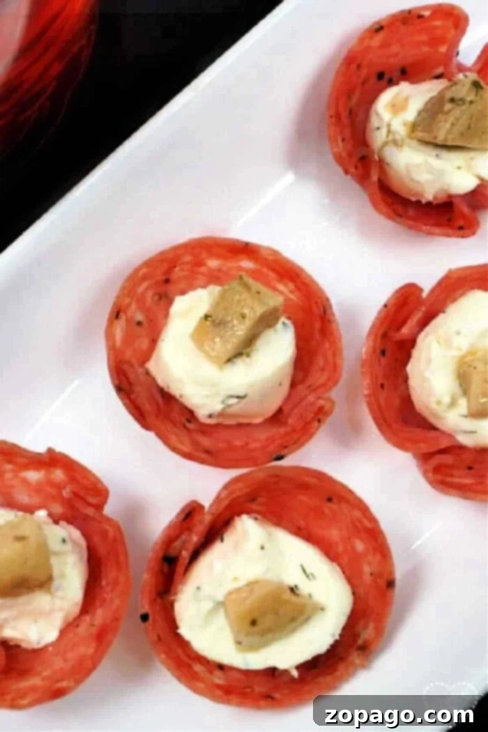 Mini Salami Bites 2 A close-up shot of a white rectangular plate featuring five perfectly arranged salami cups. Each crispy salami cup is generously filled with a creamy cheese mixture and elegantly topped with a small, marinated artichoke heart piece, making for an attractive appetizer presentation.