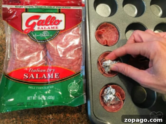 Salami Bites 3 A hand presses aluminum foil into salami slices nestled in a mini muffin tin to form savory salami cups. A package of Gallo salame rests on the counter next to the tray, indicating the main ingredient for this easy appetizer.