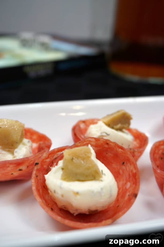 How you make salami cups! These are the best bite size cream cheese and artichoke appetizer with salami used as a little cup. You've got to bake these and serve them at your next Super Bowl party or just because friends are coming over. #salami #baked #cups #appetizer #creamcheese #artichoke #fingerfood #superbowl Salami cups filled with creamy cheese and artichoke, garnished with dill. This close-up view highlights the inviting texture and colorful presentation, making them an ideal appetizer for any gathering.