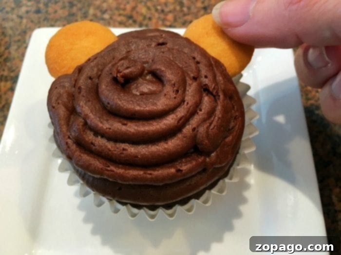 Whip Up Whimsical Monkey Cupcakes 2 A chocolate cupcake with a generous swirl of chocolate frosting, adorned with two mini vanilla wafers as ears, capturing the initial stage of crafting cute monkey cupcakes.
