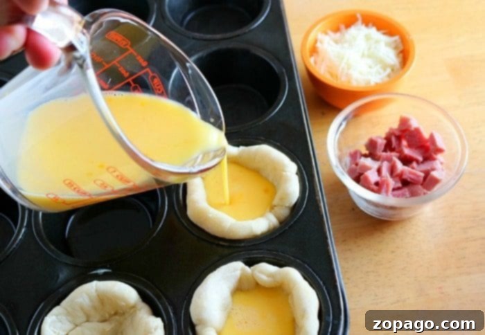 Ham and cheese breakfast bites are great using leftover ham! A great muffin tin recipe for breakfast and a perfect on the go nutritious meal kids love. #leftoverham #breakfast #muffintin #bites #biscuits A measuring cup pours egg mixture into biscuit dough cups within a muffin tin, surrounded by bowls of shredded cheese and diced ham, ready for baking.