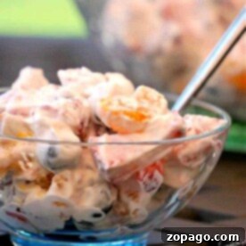 A bowl of creamy fruit salad with a spoon, featuring grapes, oranges, marshmallows, and a dollop of cool whip.