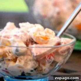 A bowl of creamy fruit salad with a spoon, featuring grapes, oranges, marshmallows, and a dollop of cool whip.