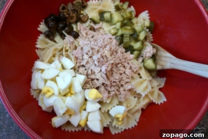 Freshly prepared macaroni tuna salad, a delightful side dish. A vibrant red bowl filled with homemade tuna macaroni salad, featuring bowtie pasta, chunks of tuna, hard-boiled eggs, diced pickles, and sliced black olives, ready to be served.