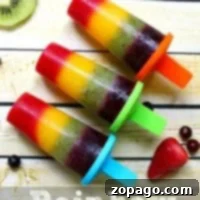 Three vibrant rainbow popsicles rest on a wooden surface, surrounded by an array of fresh fruit slices, showcasing their appealing colors.