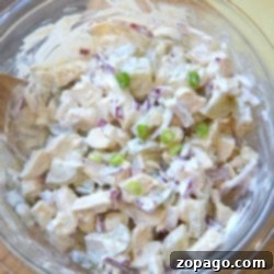 The Ultimate Chicken Salad Sandwich Recipe 6 A glass bowl brimming with creamy rotisserie chicken salad, featuring diced onions, pickles, and a medley of flavors sits enticingly on a wooden board.