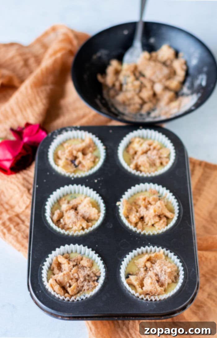 Make-Ahead Blueberry Muffins 7 A muffin tray with six unbaked blueberry muffins with frozen blueberries sits next to a bowl of crumb topping and a red rose on a brown cloth.