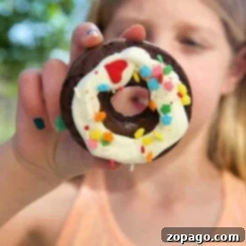 A child gleefully holds a chocolate donut crafted from cake mix, topped with white icing and vibrant, colorful sprinkles.
