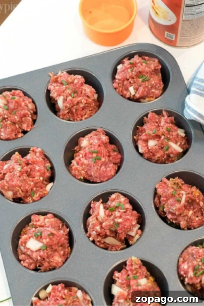 Mini Meatloaf Muffins 2 A muffin tin filled with raw meatloaf mixture ready to be baked. The meatloaf muffin recipe portions appear roughly shaped and are topped with bits of onion and herbs. An orange bowl and a tin can are visible in the background.