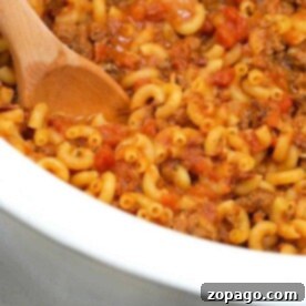 A wooden spoon stirs a pot of macaroni with ground meat and tomato sauce, reminiscent of a homemade crockpot hamburger helper.