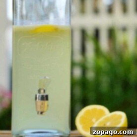 A glass container of fresh squeezed lemonade sits with a spout, accompanied by two lemon halves on a wooden surface, offering a glimpse into the perfect summer recipe.
