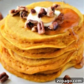 A stack of pumpkin pancakes, one of the easy pumpkin desserts, topped with butter, pecans, and drizzled maple syrup sits on a plate. The pancakes have a light golden-brown color.