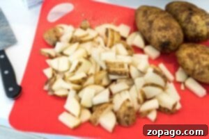 Creamy Slow Cooker Mashed Potatoes 4 Diced russet potatoes on a red cutting board next to whole potatoes and a knife, ready for slow cooking.