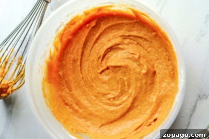 Simple Pumpkin Bundt Bites 3 A bowl of creamy pumpkin cake mix with a whisk sits on a white marble countertop, ready to transform into delightful mini bundt cakes.