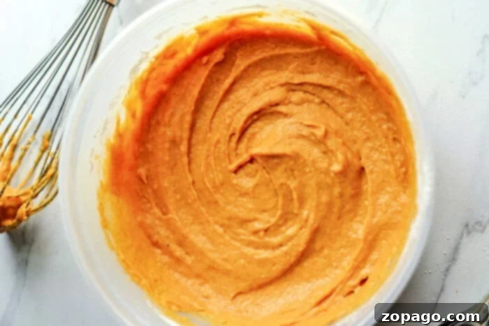 Easy Pumpkin Bundtlets 3 A bowl of creamy pumpkin cake mix with a whisk sits on a white marble countertop, ready to transform into delightful mini bundt cakes.