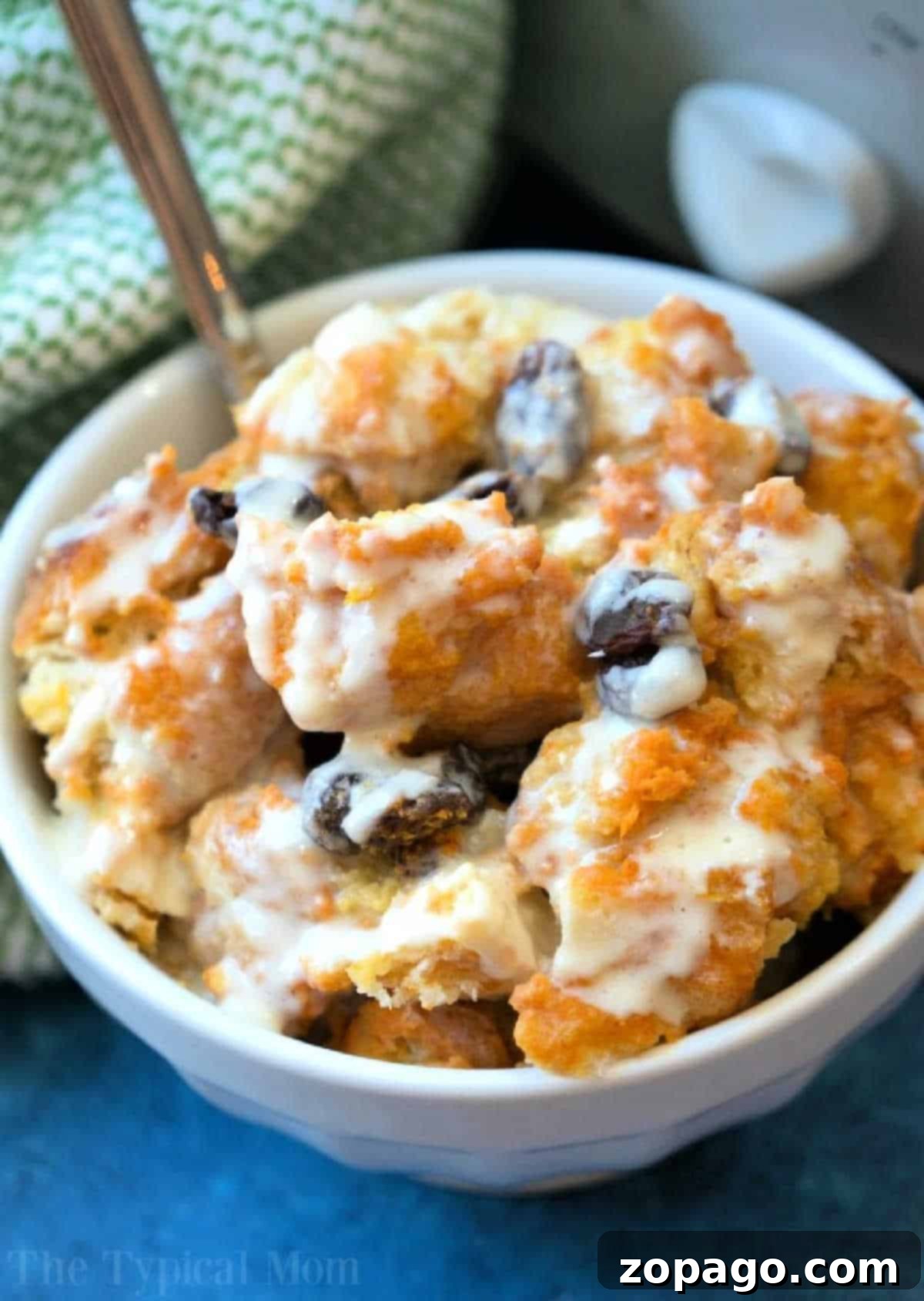 A bowl of sweet crockpot bread pudding with raisins, topped with creamy icing or sauce, sits beside a green patterned cloth, inviting a delightful experience.