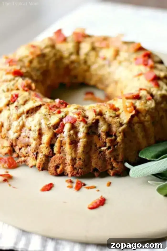 Mastering Stuffing in a Bundt Pan 5 A savory bundt cake, shaped perfectly by the bundt pan, boasts a golden-brown crust and is topped with crispy bacon bits. Fresh sage leaves are artfully placed beside it on a beige surface, enhancing its rustic charm.