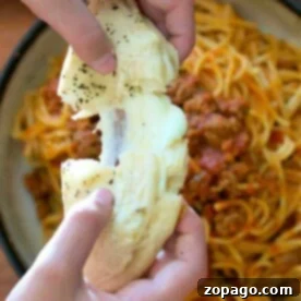 Hands pulling apart cheesy garlic bread over a plate of spaghetti with meat sauce, using flaky layers reminiscent of a refrigerated biscuit filled with cheese.