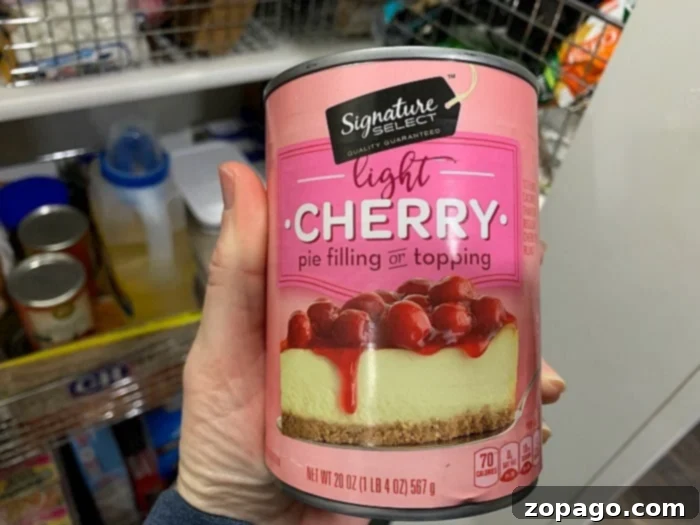 Inverted Cherry Delight 2 A hand holds a can of Signature Select light cherry pie filling, highlighting its convenience for quick desserts. The label shows cherries on a cheesecake, while a blurred background displays other grocery items.