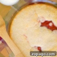 A delectable upside down cherry pie in a glass dish showcases its visible red filling beneath a golden crust. A wooden spatula rests on a plaid cloth, while a glass of milk stands invitingly in the background.