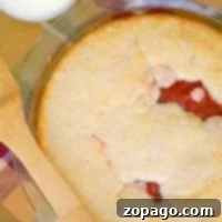 A delectable upside down cherry pie in a glass dish showcases its visible red filling beneath a golden crust. A wooden spatula rests on a plaid cloth, while a glass of milk stands invitingly in the background.