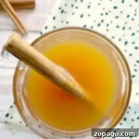 Top view of a crockpot hot apple cider in a glass, adorned with a cinnamon stick. The drink rests on a wooden surface beside a white cloth with green dots, while another cinnamon stick peeks onto the table.