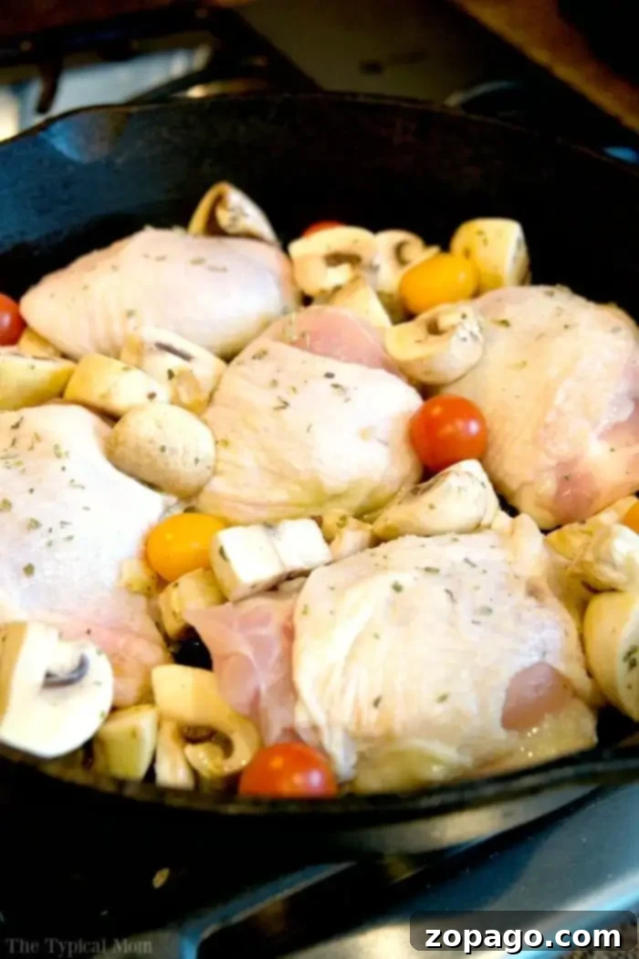 Baked bone in chicken thighs recipe that is made in one pot! So easy and done from start to finish in just 30 minutes with only 5 ingredients. Delicious. A cast iron skillet on a stove contains raw chicken thighs, quartered mushrooms, and cherry tomatoes, ready for cooking. While you prep your ingredients, you might wonder, *How long to bake bone-in chicken thighs at 350*? Seasoned with herbs, this dish promises a flavorful experience.