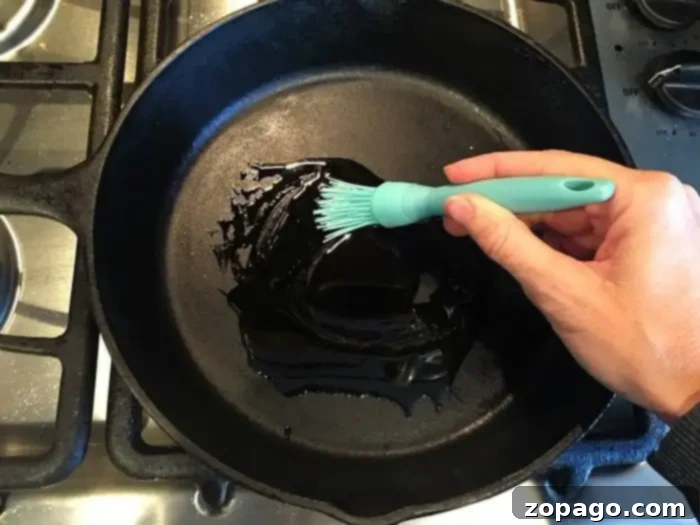 Mastering Bone-in Chicken Thighs: The Optimal Baking Time 2 A hand holding a blue silicone brush is spreading oil on a black cast-iron skillet, preparing it for cooking. The skillet is placed on a stove with metal burners and control knobs visible on the side. Meanwhile, wondering how long to bake bone-in chicken thighs at 350 can ensure perfectly cooked results.