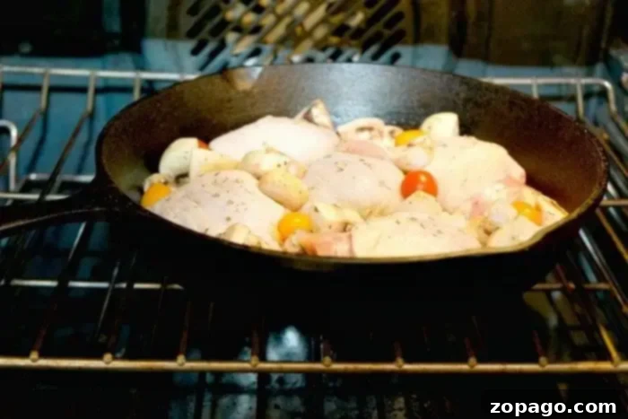 Mastering Bone-in Chicken Thighs: The Optimal Baking Time 3 A cast iron skillet containing raw chicken pieces, cherry tomatoes, and mushrooms is in an oven with the door closed. The oven's heating elements are visible, and the food appears to be seasoned with herbs. For those wondering how long to bake bone-in chicken thighs at 350, this setup offers a savory answer.